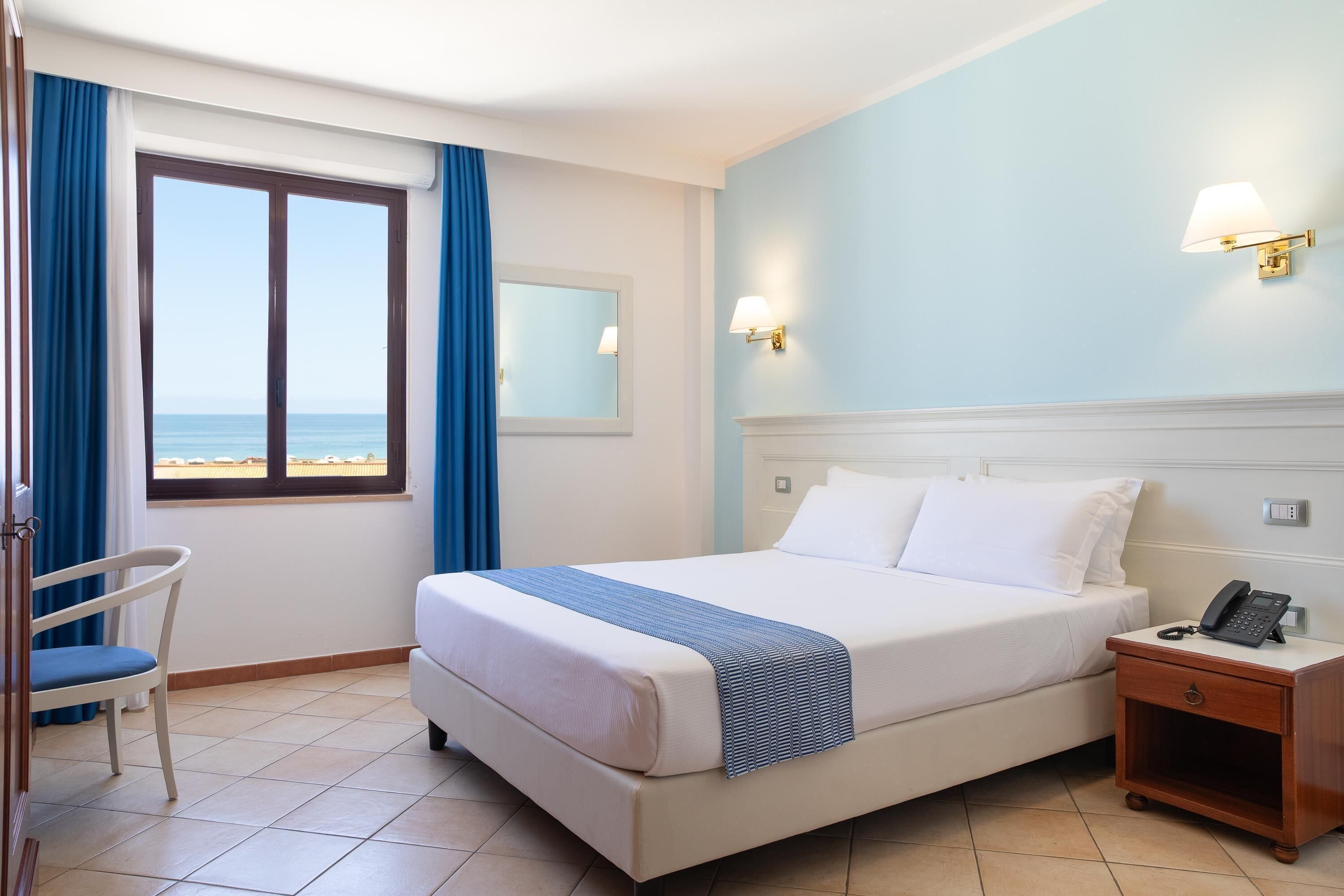 A hotel room with a bed, bedside table, chair, and a window with a view of the sea.