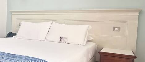 Una cama bien hecha con ropa blanca y almohadas, una mesita de noche con lámpara y un enchufe de pared.