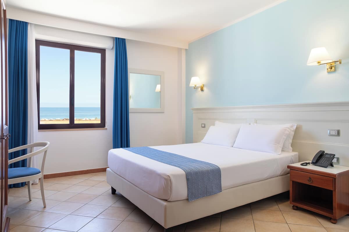 Una camera d'albergo con un letto, un comodino con il telefono, una sedia, una finestra con tende e una lampada a parete.
