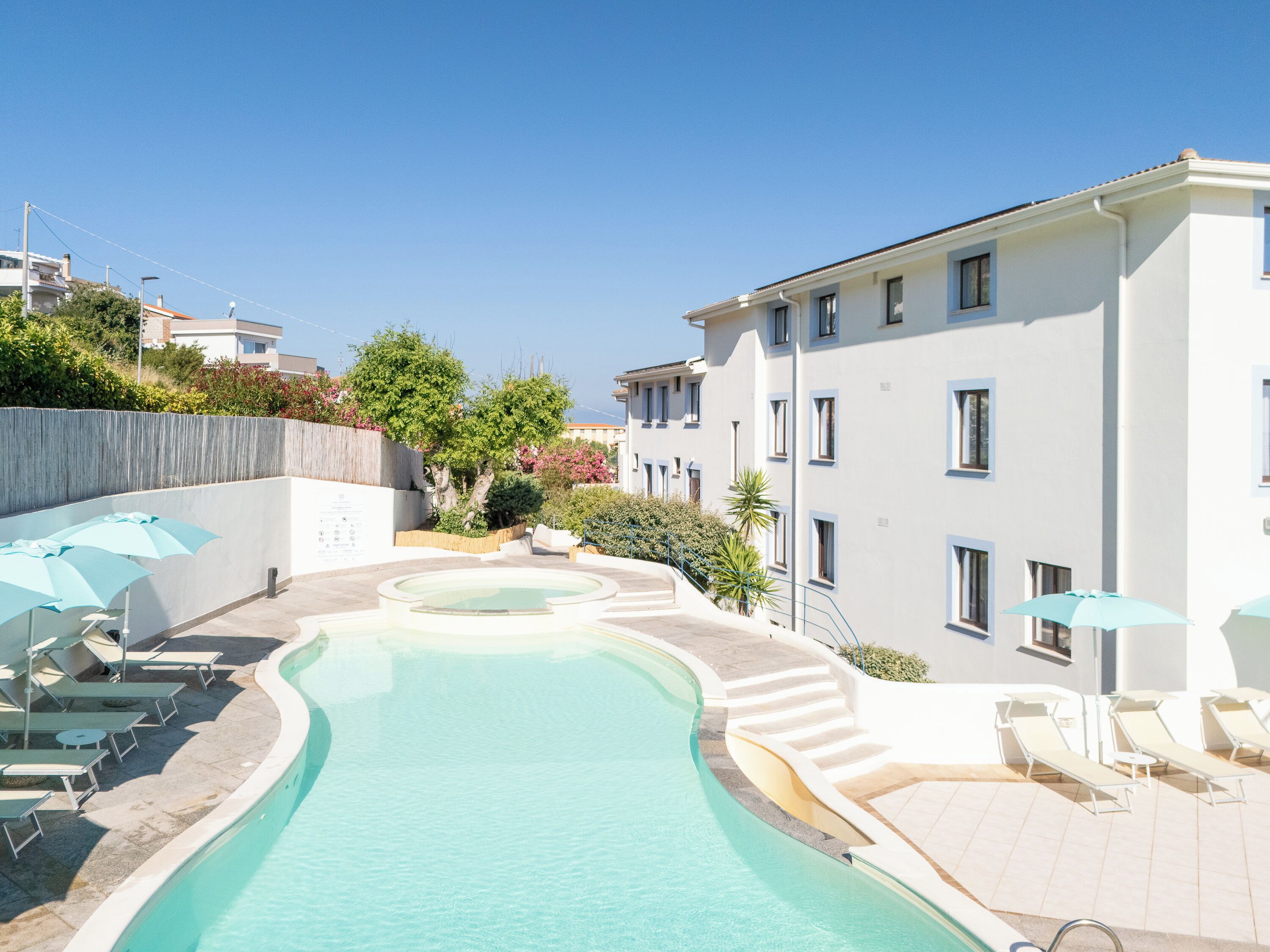 Ein Schwimmbecken mit Liegestühlen und Sonnenschirmen, umgeben von einem weißen Gebäude mit mehreren Fenstern und einer Treppe, die zu ihm hinaufführt.