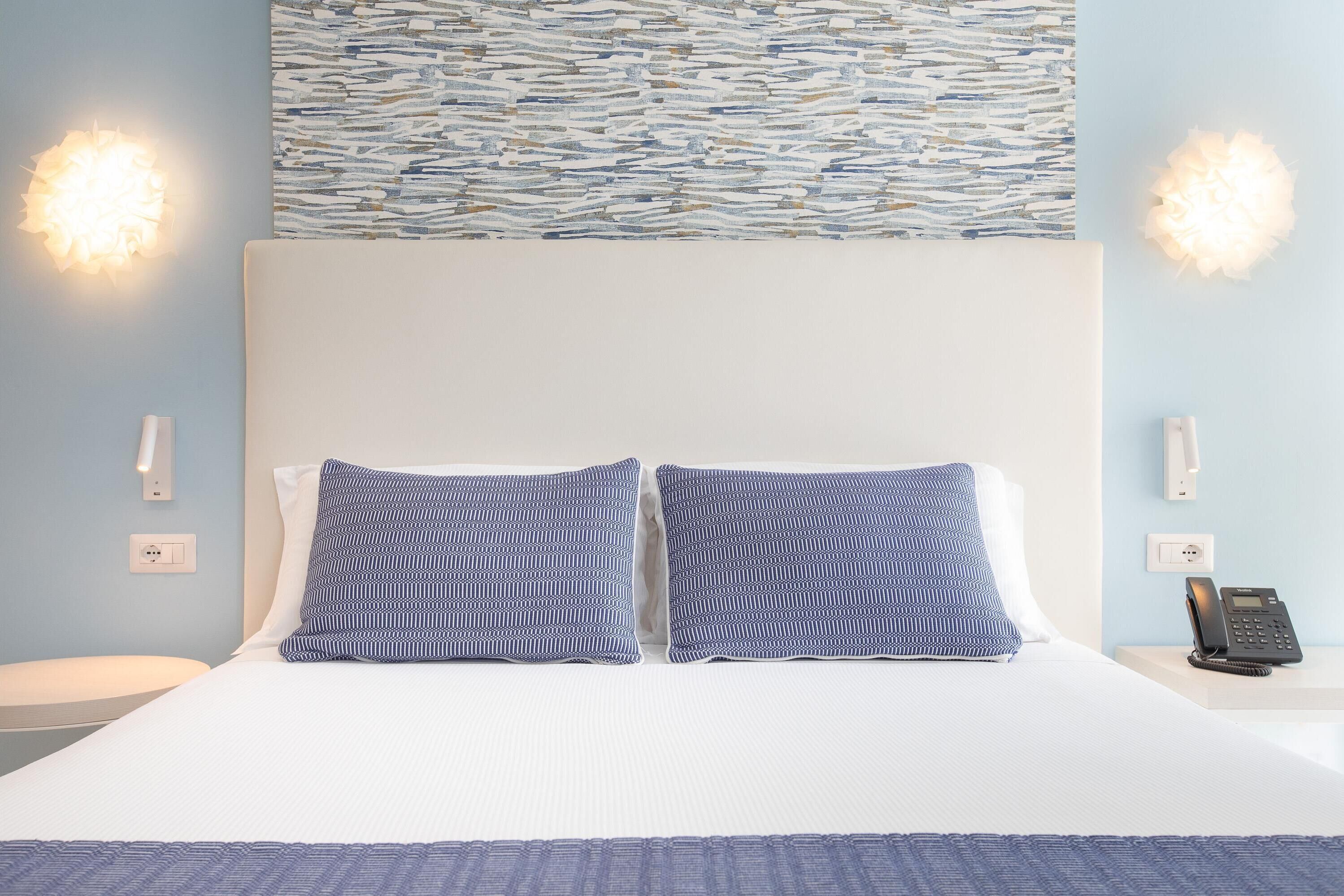 Quarto de hotel com cama grande, duas luminárias de cabeceira, um telefone na mesinha de apoio e uma obra de arte com textura na parede acima da cama.