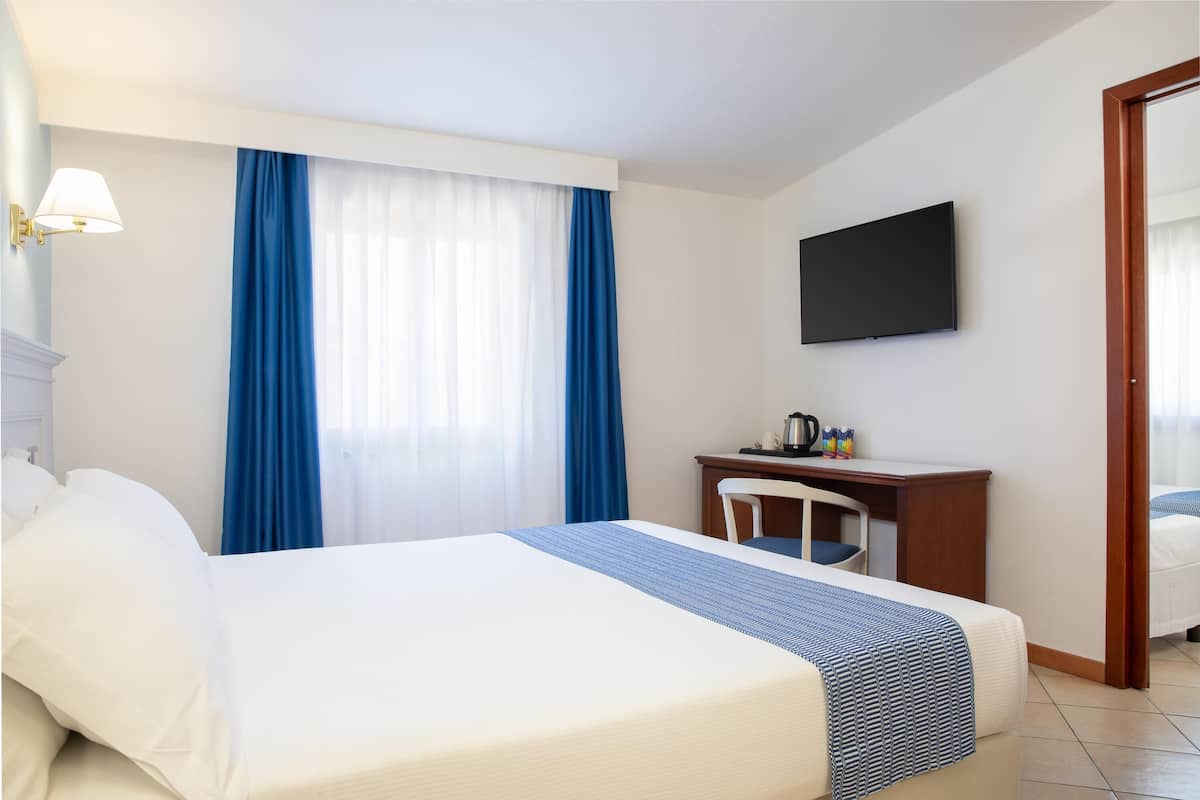 Una camera d'albergo con un letto, una scrivania, una sedia, una TV a schermo piatto e una finestra con tende blu.
