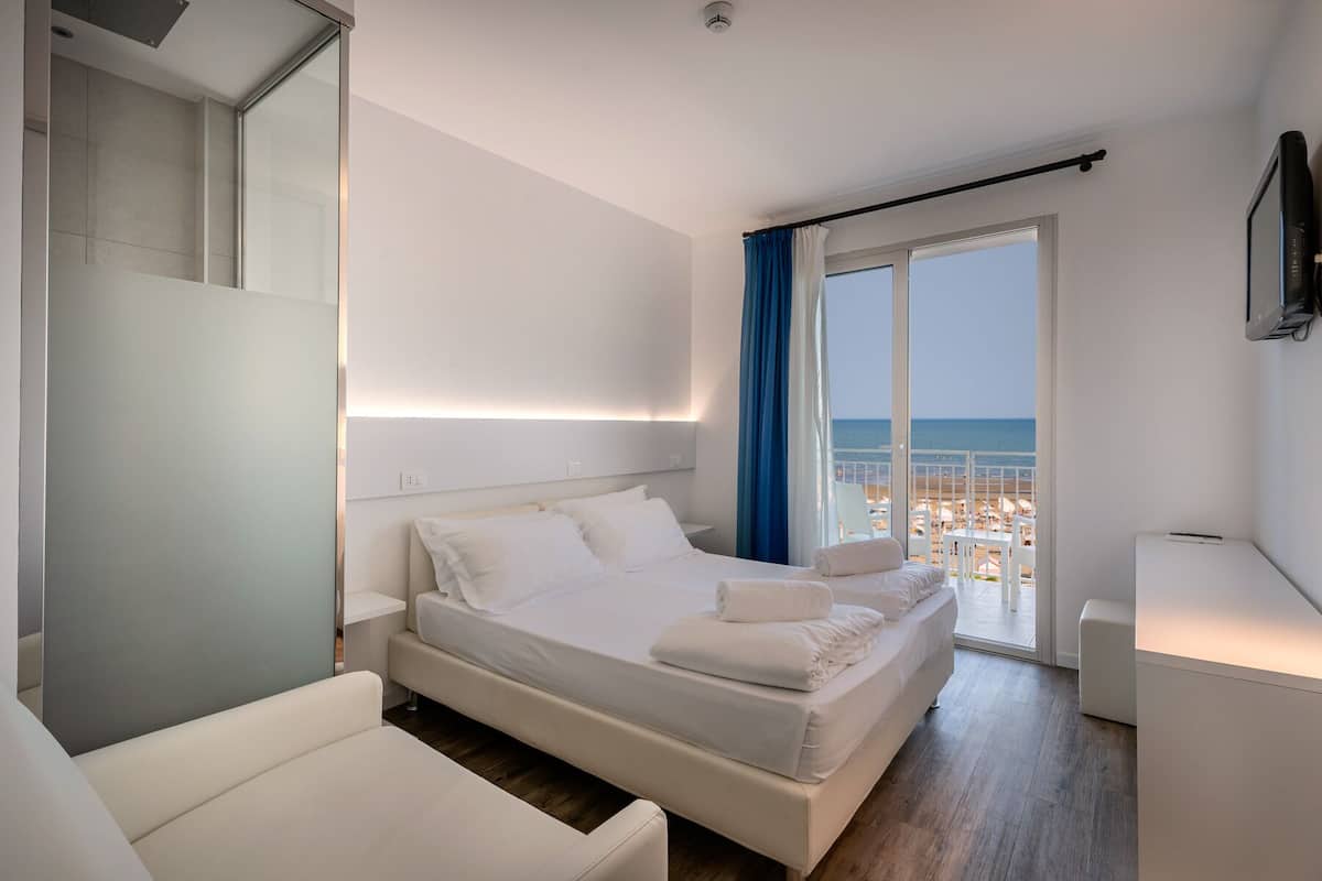 A modern hotel room with a large bed, a seating area, and a balcony with a view of the beach.