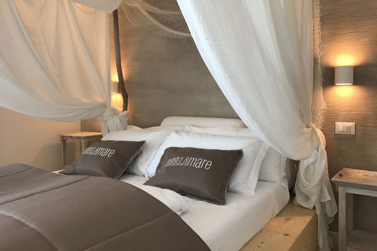A bed with white bedding and two pillows with "STELLA MARE" text, a canopy, a bedside table, and a wall-mounted light.