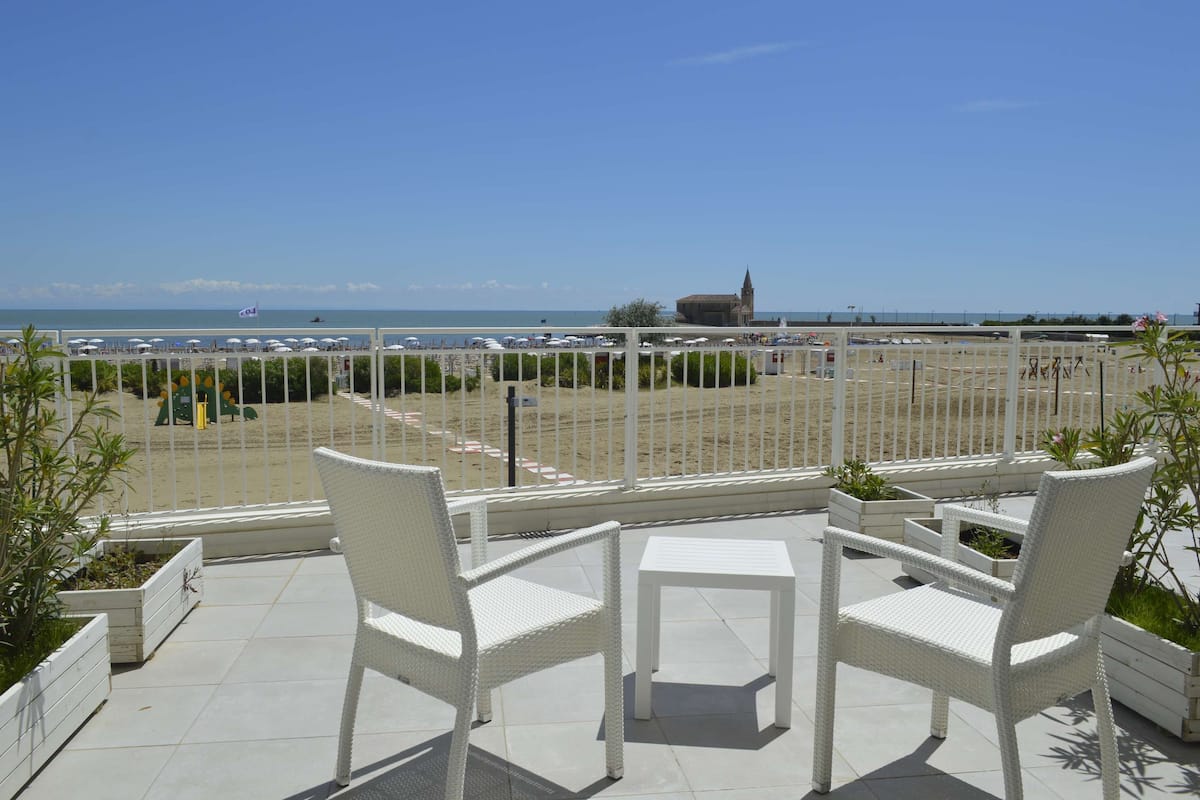 A balcony with white wicker furniture, a view of the beach, and a distant church.