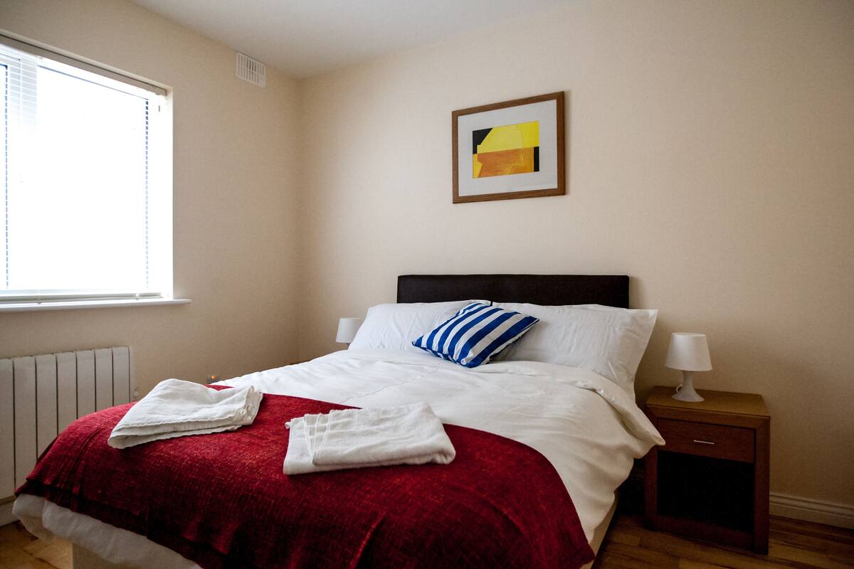 A neatly made bed with a red blanket, white linens, and a blue-striped pillow. There is a small bedside table with a lamp and a framed picture on the wall above the bed.