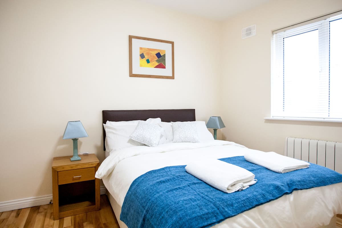 A neatly made bed with white linens and a blue blanket, a small bedside table with a lamp, and a window with blinds.