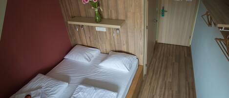 Una cama bien hecha con sábanas y almohadas blancas, un cabecero de madera, un pequeño estante con un florero y una puerta de madera con pomo.