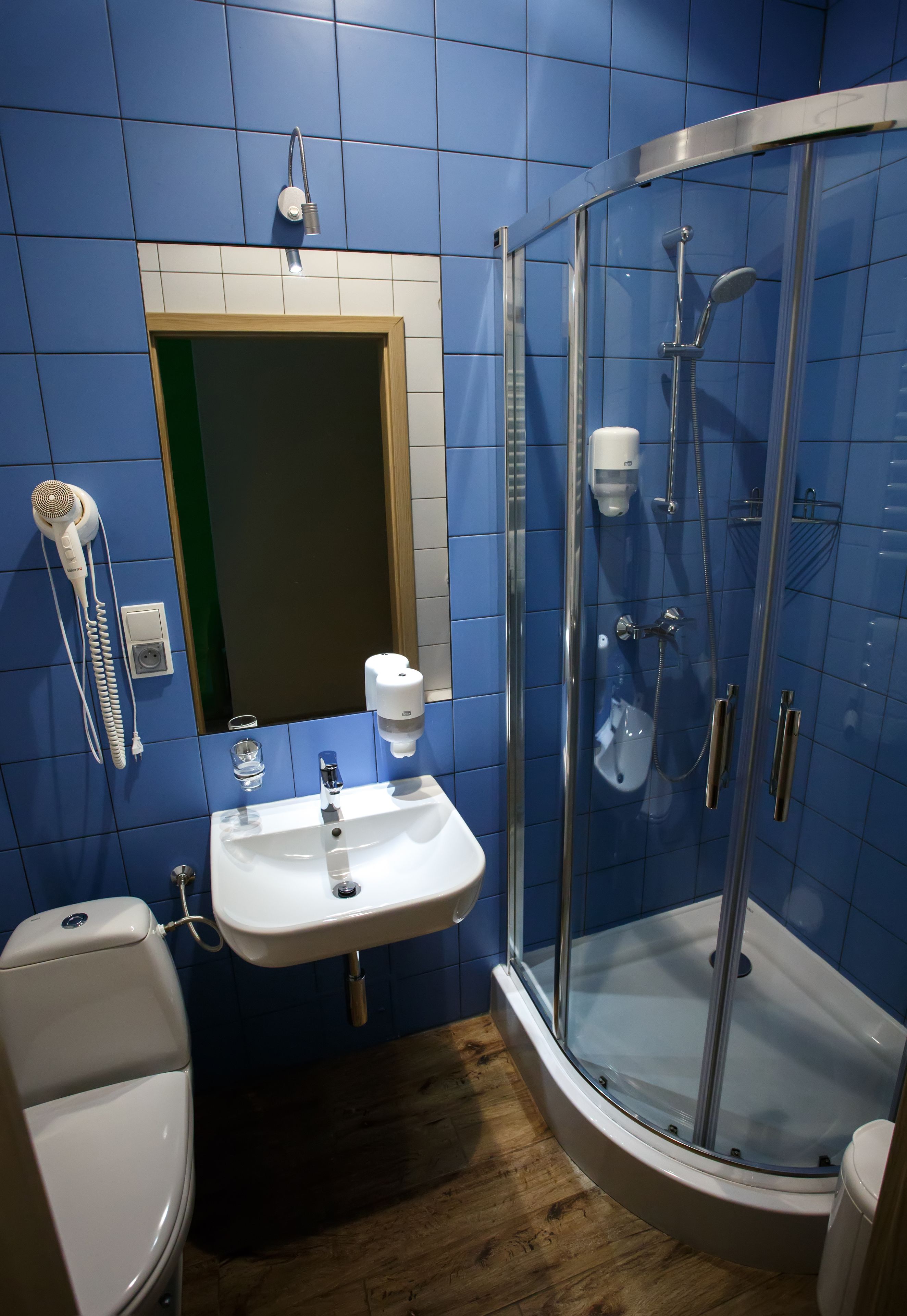 Ein Badezimmer mit blauen Fliesen, einem weißen Waschbecken, einer Duschkabine aus Glas und einem an der Wand befestigten Föhn.