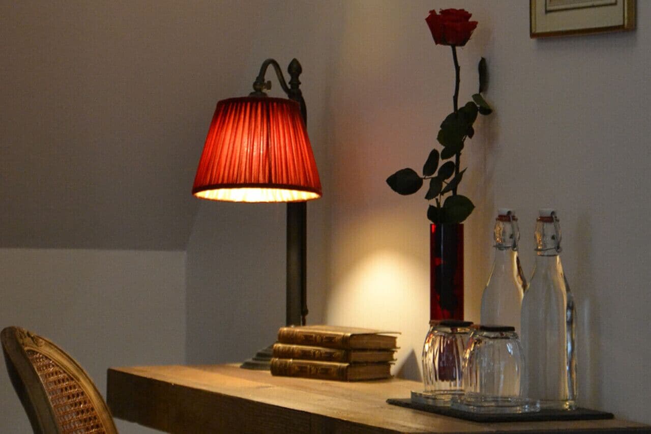 A table with a lamp, books, a vase with a rose, and glass bottles.