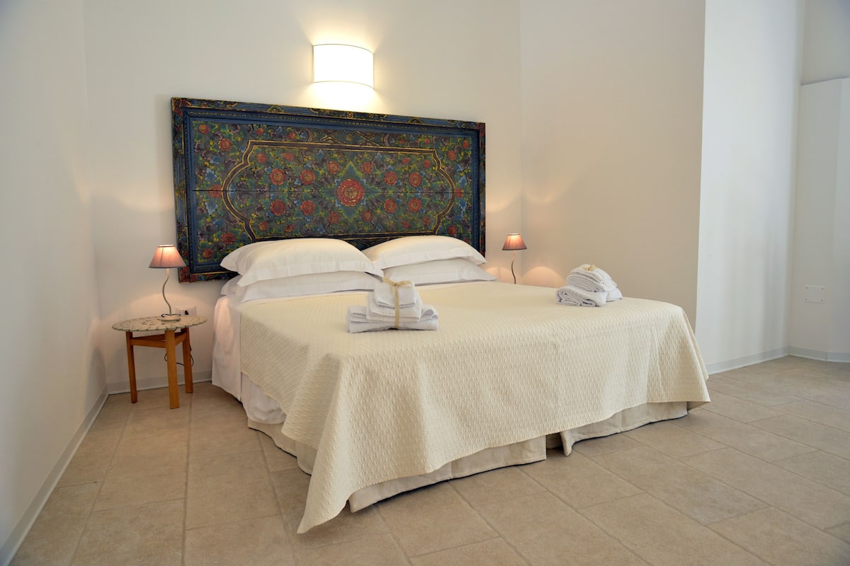 Una cama bien hecha con un tablero de cabecera decorativo, dos mesitas de noche con lámparas y una mesita redonda pequeña con una lámpara.