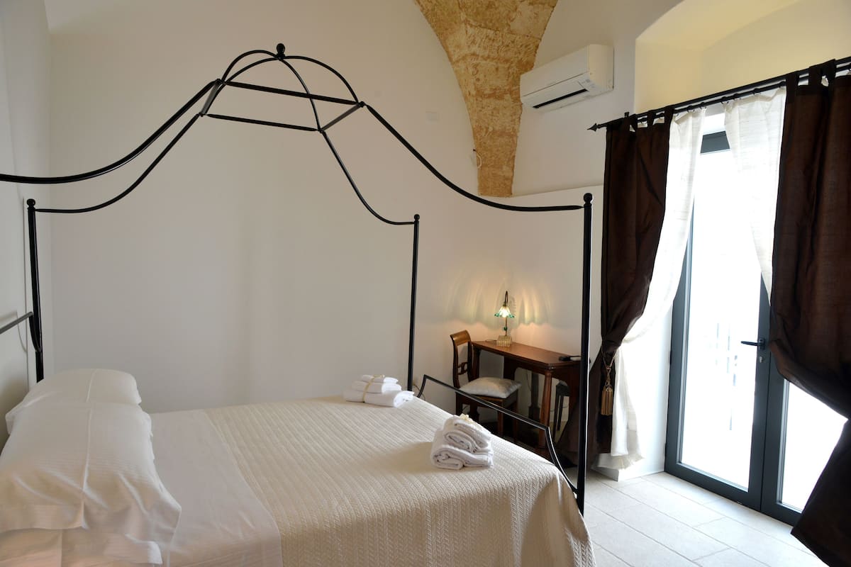 Una cama bien tendida con sábanas blancas, una silla de madera, una mesita con una lámpara y una ventana con cortinas.