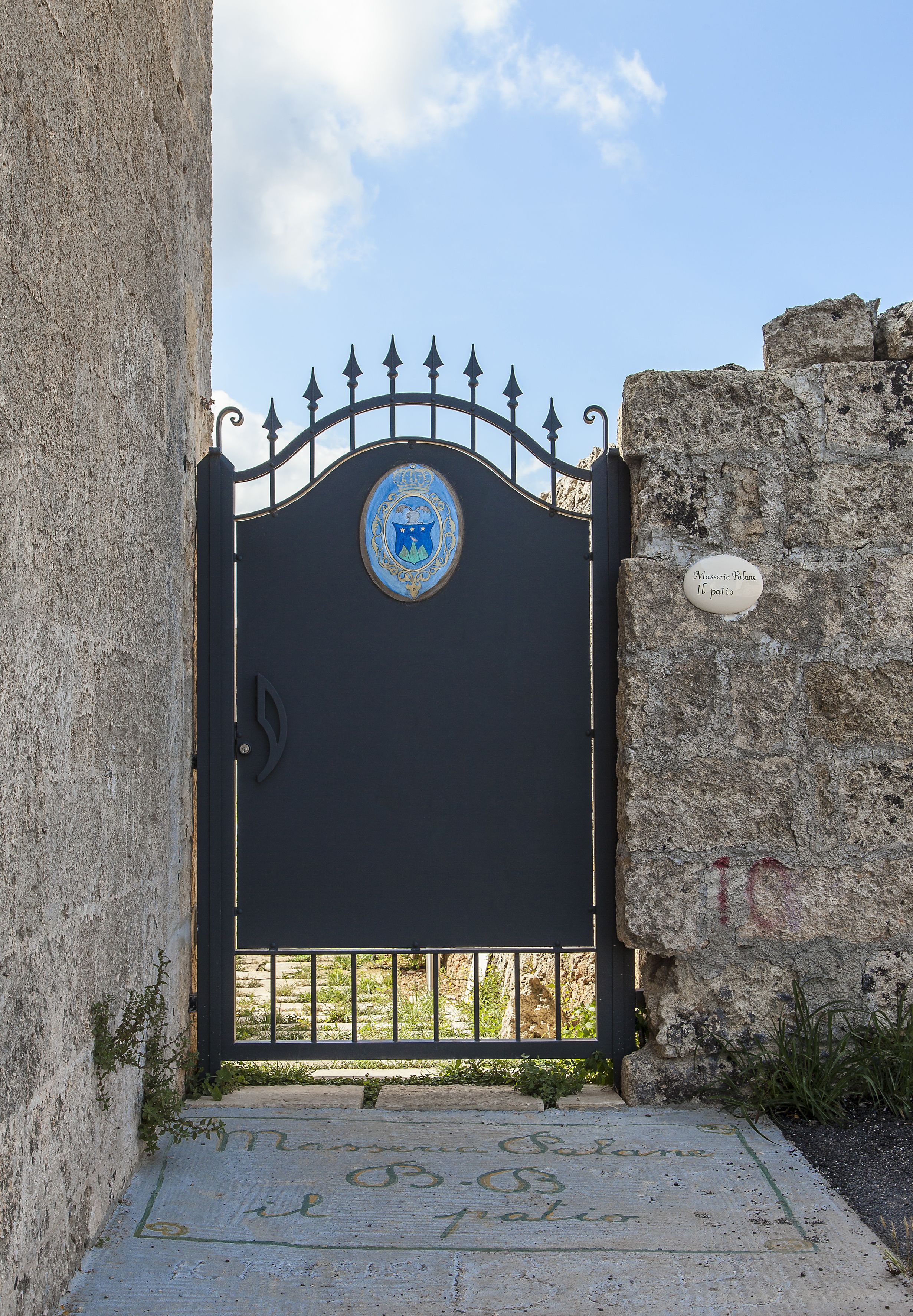 A black metal gate with a decorative emblem, a stone wall, and a sign reading "Mansion Palace Il palio".