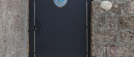 A black metal gate with a decorative emblem, a stone wall, and a sign reading "Mansion Palace Il palio".