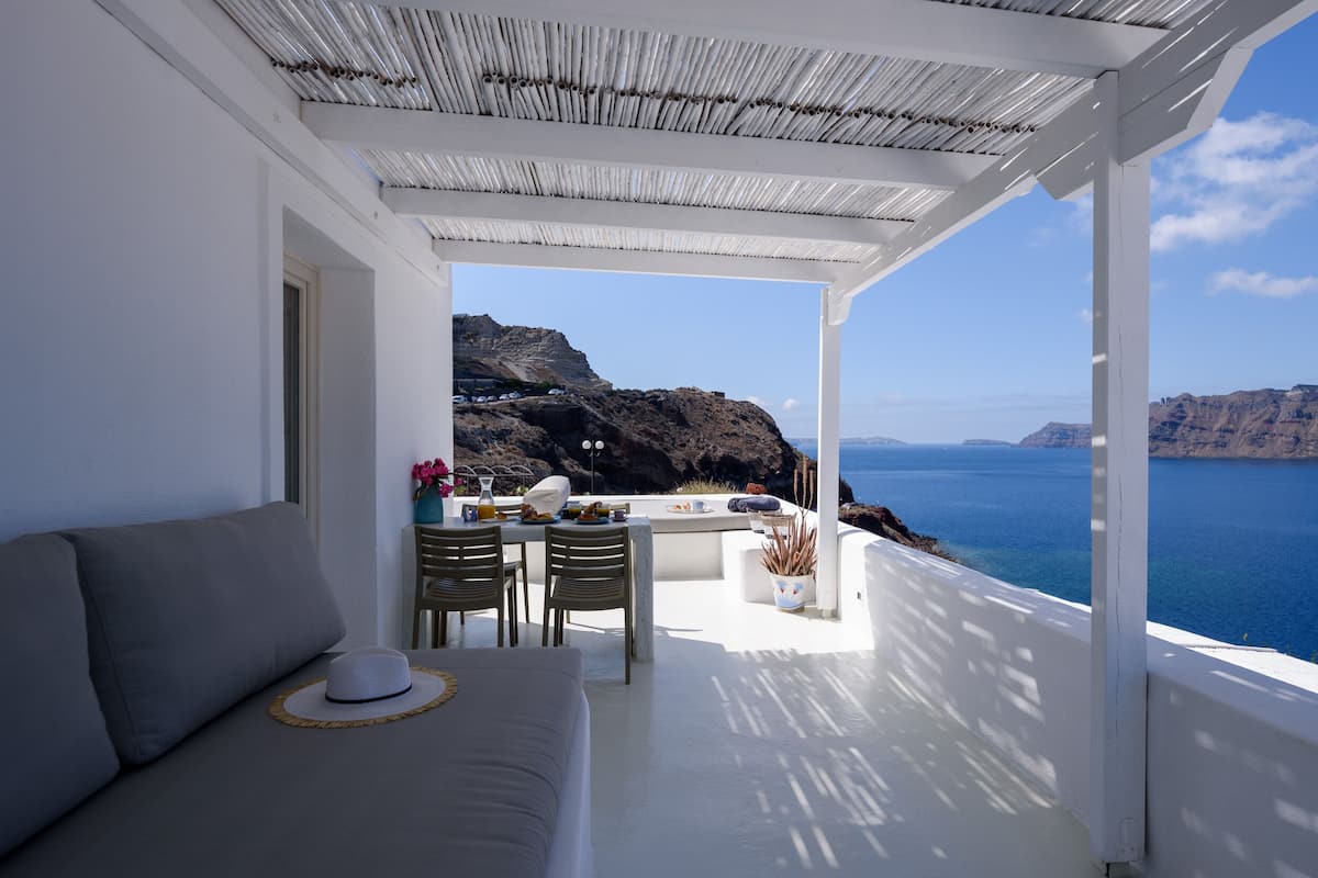 A balcony with a white railing, a sofa, and a table with chairs. The view includes a body of water and rocky cliffs.