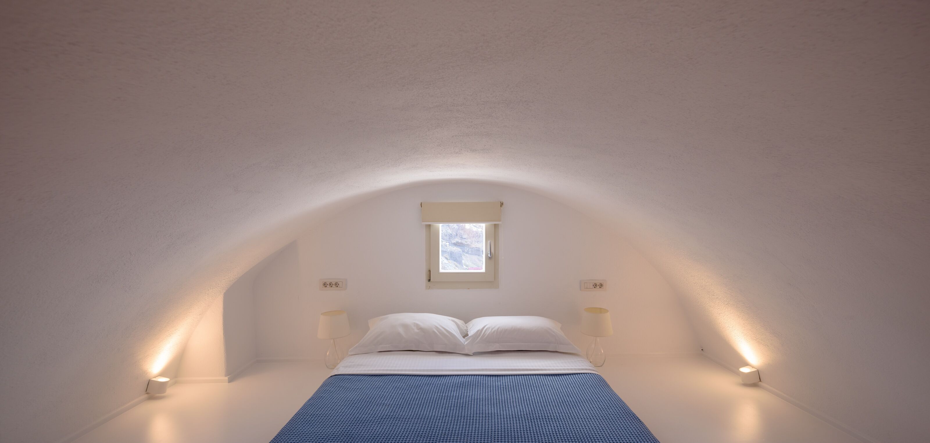A bedroom with a sloped ceiling, a single bed, two bedside lamps, and a window with a white blind.