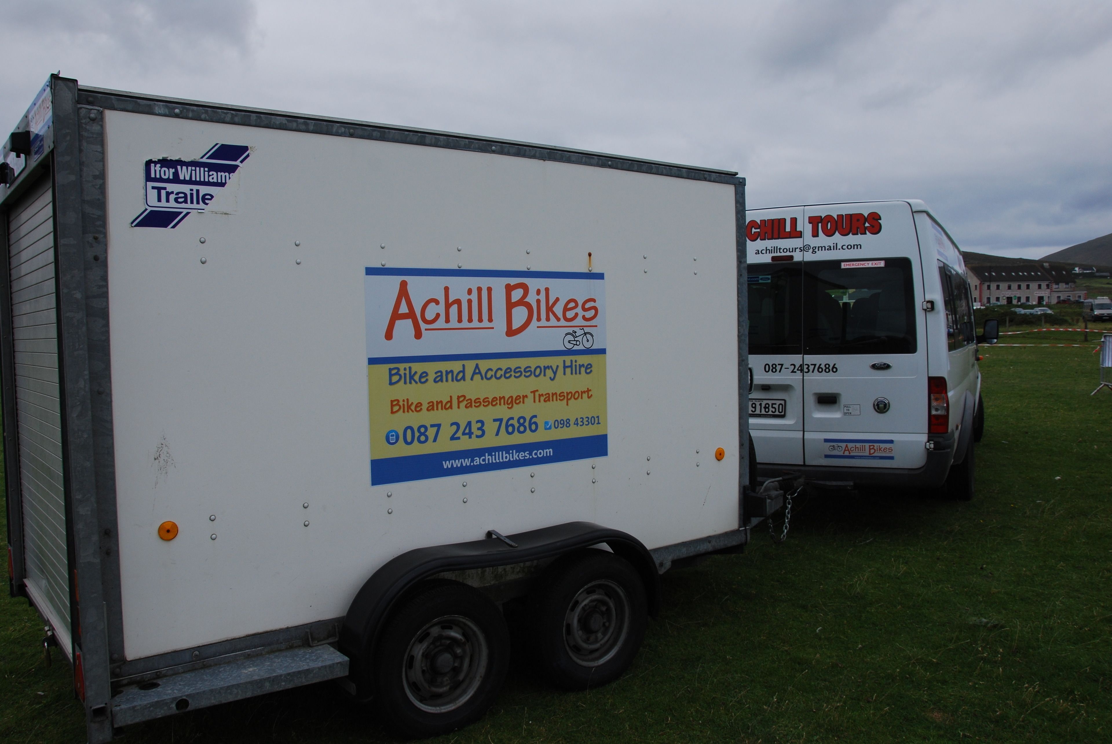 Un remolque blanco con la marca "Achill Bikes", que incluye información de contacto y servicios como transporte de bicicletas y pasajeros.