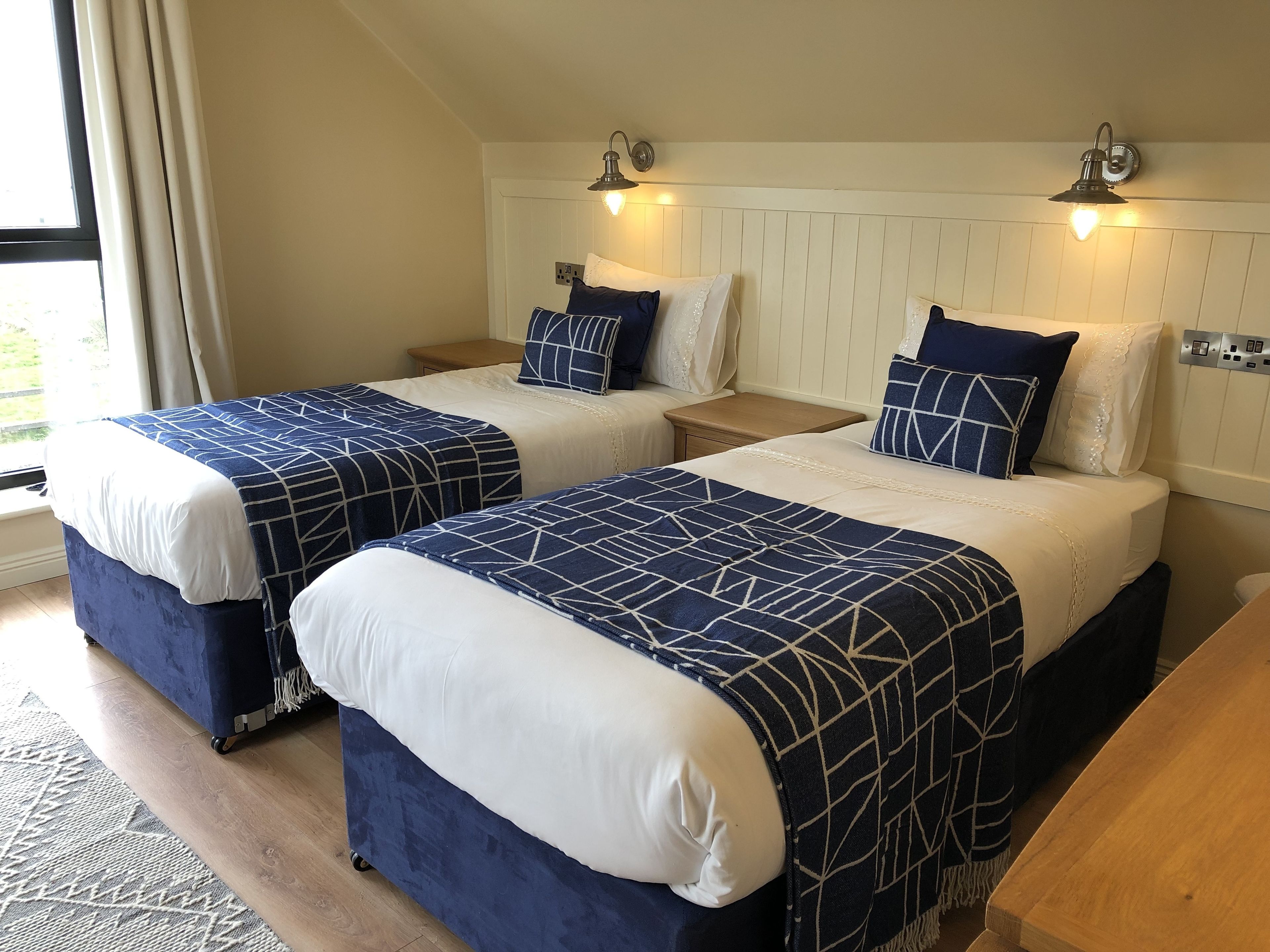 Ein Schlafzimmer mit zwei Betten, jedes mit einer blau-weißen Bettdecke, einem hölzernen Nachttisch und einer Wandleuchte.