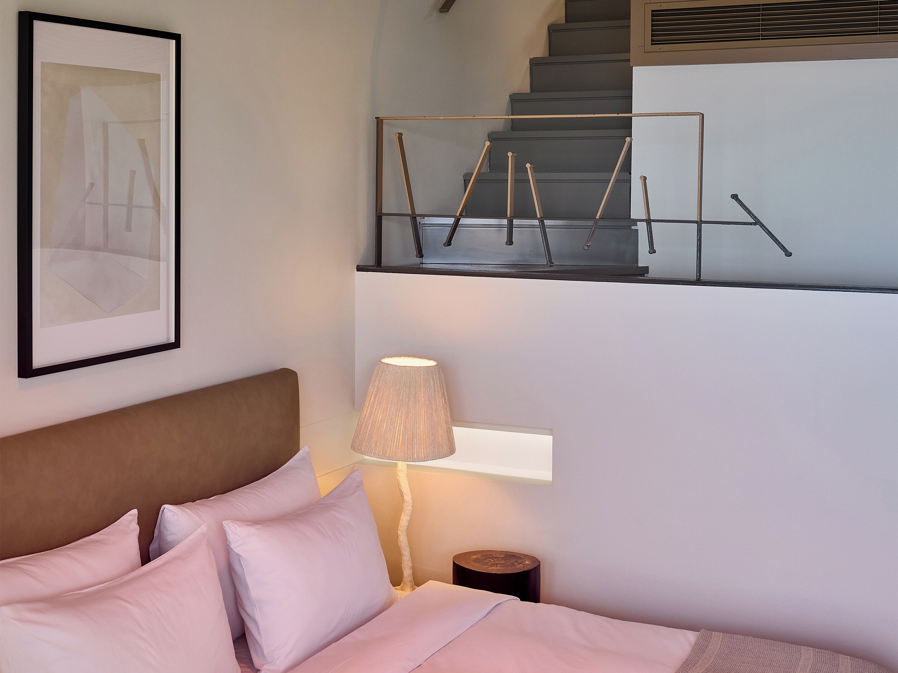 Une chambre à coucher avec un lit, des oreillers, une lampe de chevet, un tableau encadré et un escalier.