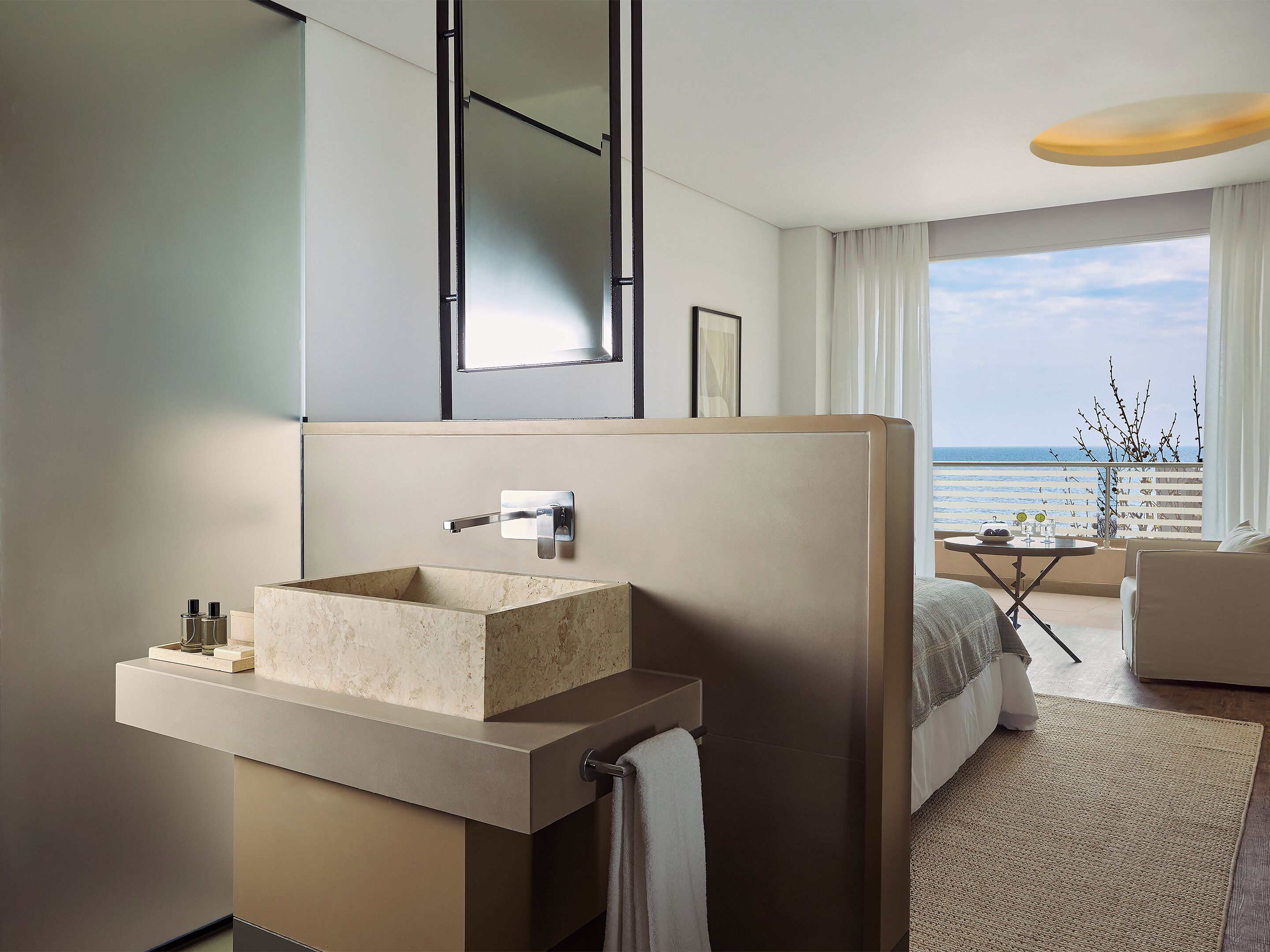 A modern hotel room with a large window, a built-in sink, a bed, and a small table with a view of the sea.