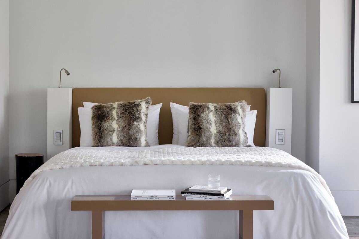 A neatly made bed with a white duvet, two pillows with a fur-like pattern, and a bedside table with a few books and a glass of water.