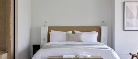 A neatly made bed with white bedding and a wooden headboard, a small bedside table with books, and a framed abstract painting on the wall.