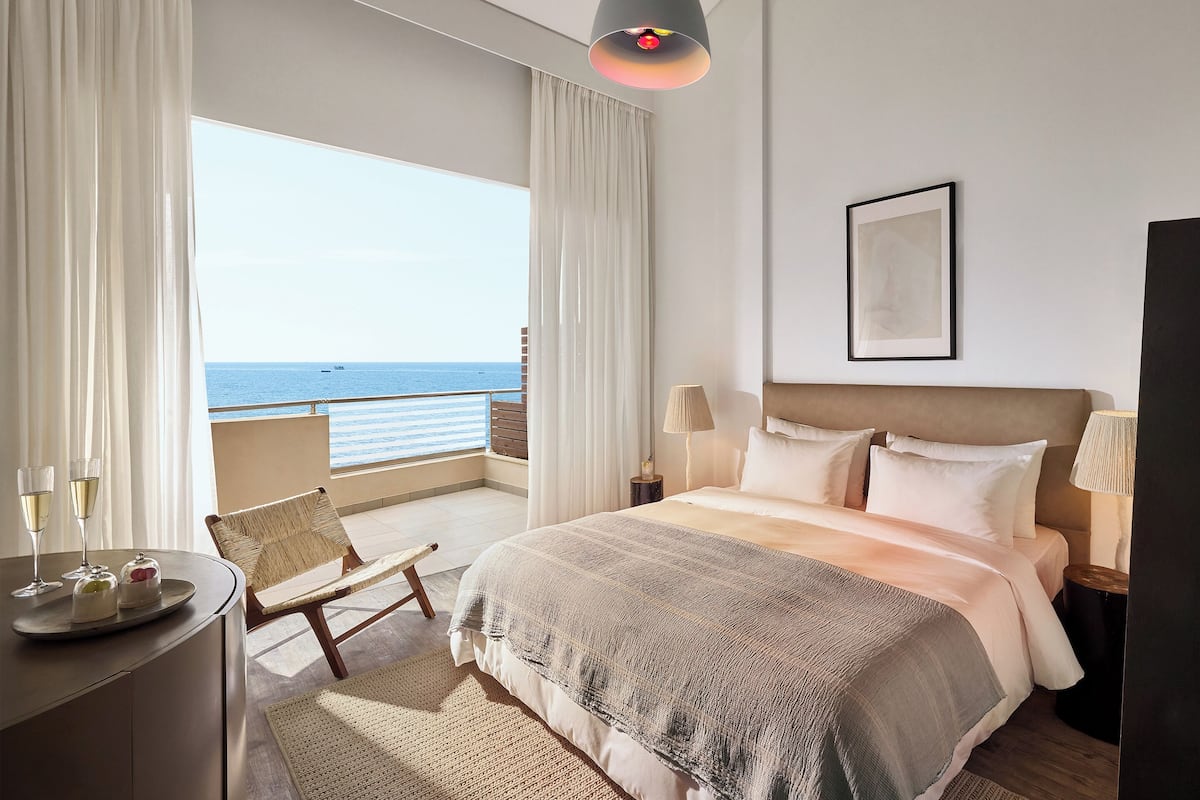 A hotel room with a bed, a desk with a chair, a lamp, and a view of the sea.