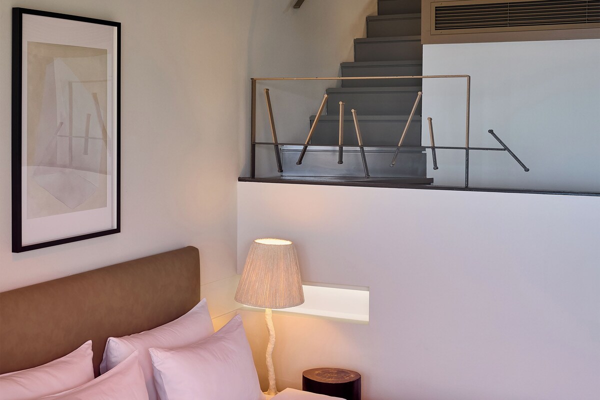 Un dormitorio con cama, almohadas, lámpara de noche, una obra de arte enmarcada y una escalera.