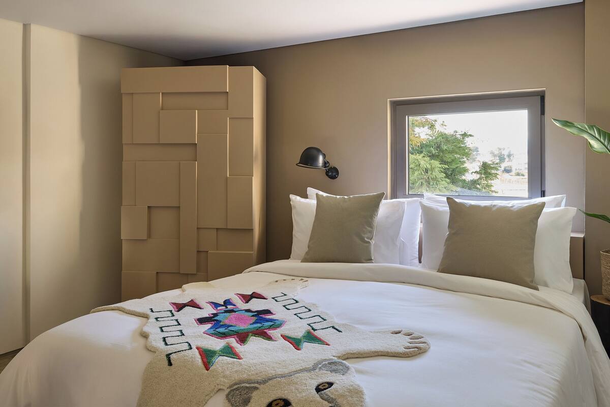 A bedroom with a bed, pillows, a wall-mounted lamp, a window with a view, and a door with a geometric design.