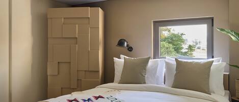 Una cama bien hecha con una manta decorativa, una lámpara de pared y una ventana con vistas a la vegetación.