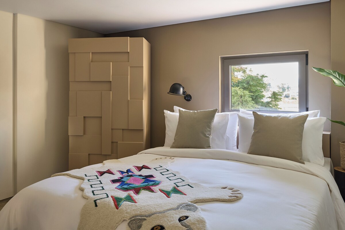 A neatly made bed with a decorative blanket, a wall-mounted lamp, and a window with a view of greenery.