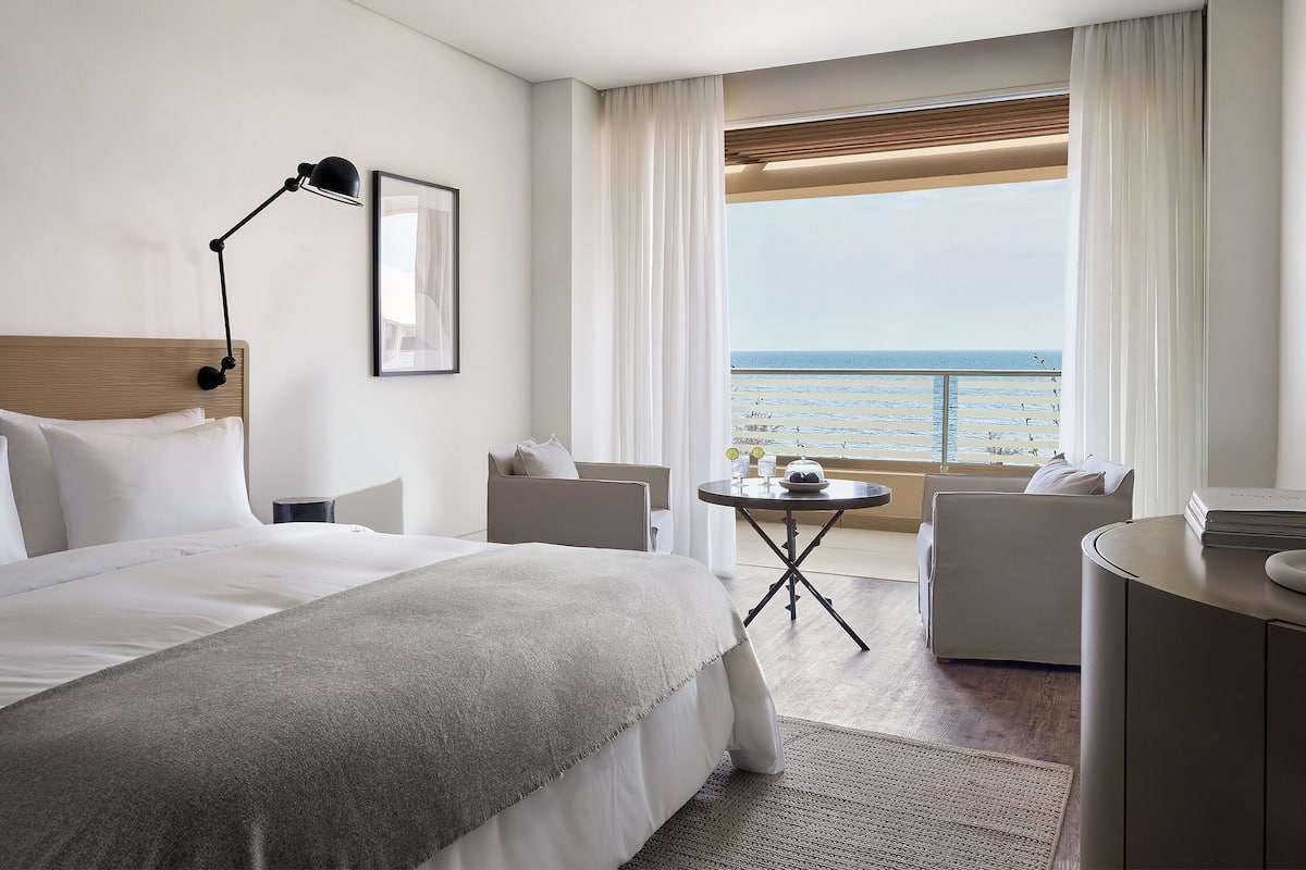 A hotel room with a bed, a desk, a chair, a small table, and a view of the sea.
