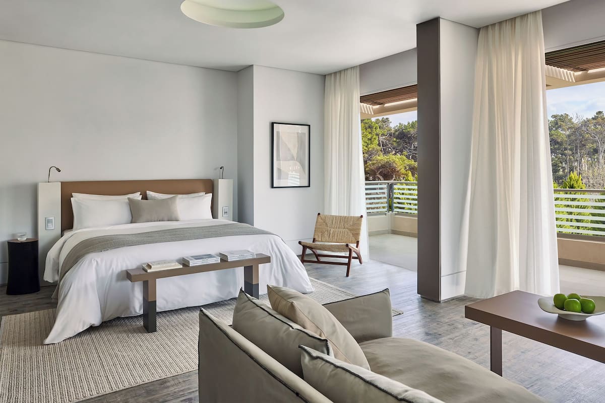 A modern hotel room with a large bed, a sitting area with a sofa and a table, and a balcony with a view of trees.