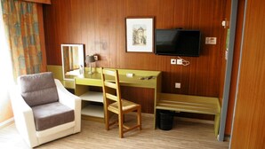 A hotel room with a wooden paneled wall, a desk with a chair, a television, a framed picture, and a window with curtains. - Le Skippy (Saint-Pierre-des-Corps)