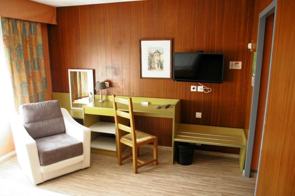A hotel room with a wooden paneled wall, a desk with a chair, a television, a framed picture, and a window with curtains.