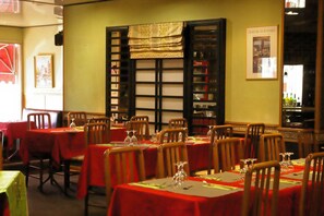 A restaurant with multiple tables covered with red tablecloths, wooden chairs, and a bookshelf in the background.