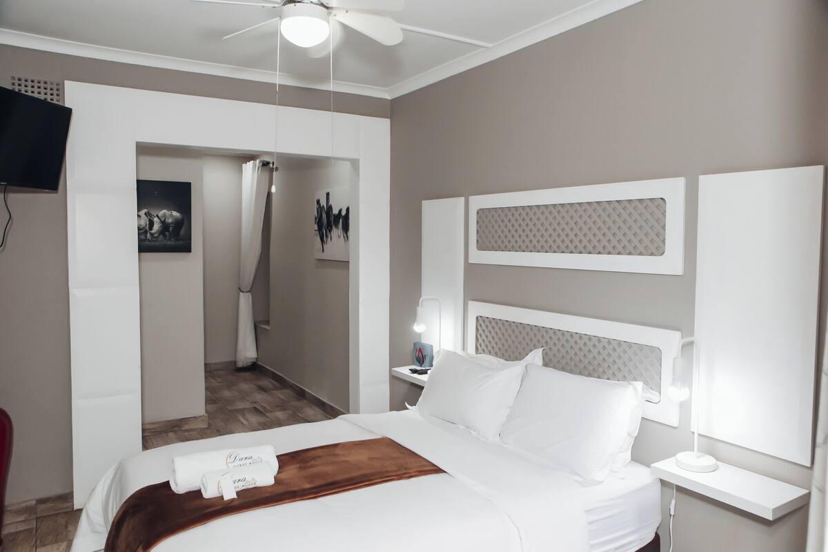 Una habitación de hotel con cama, mesita de noche, ventilador de techo, televisor montado en la pared y puerta que da a un pasillo.