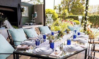 Un área de comedor al aire libre con una mesa de vidrio dispuesta para seis personas, rodeada de sillas acolchadas en azul claro.