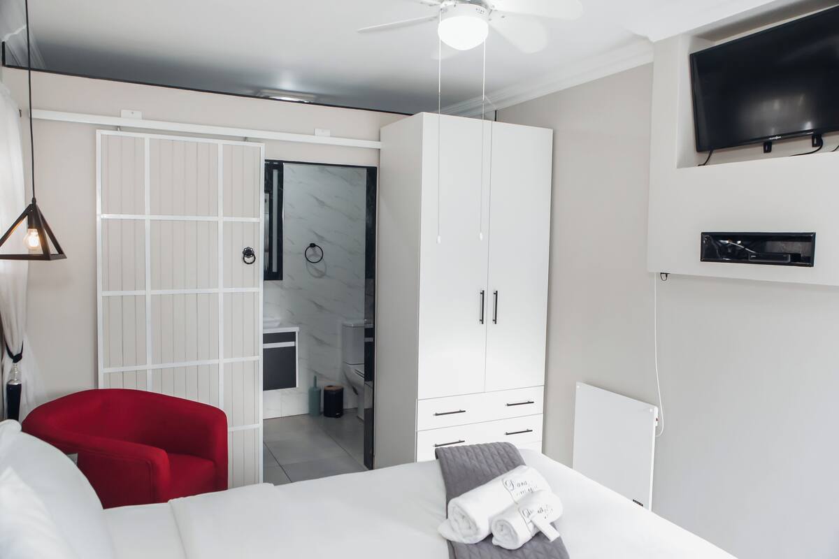 Una habitación de hotel con cama, una silla roja, un armario, televisor y un baño visible a través de una puerta abierta.