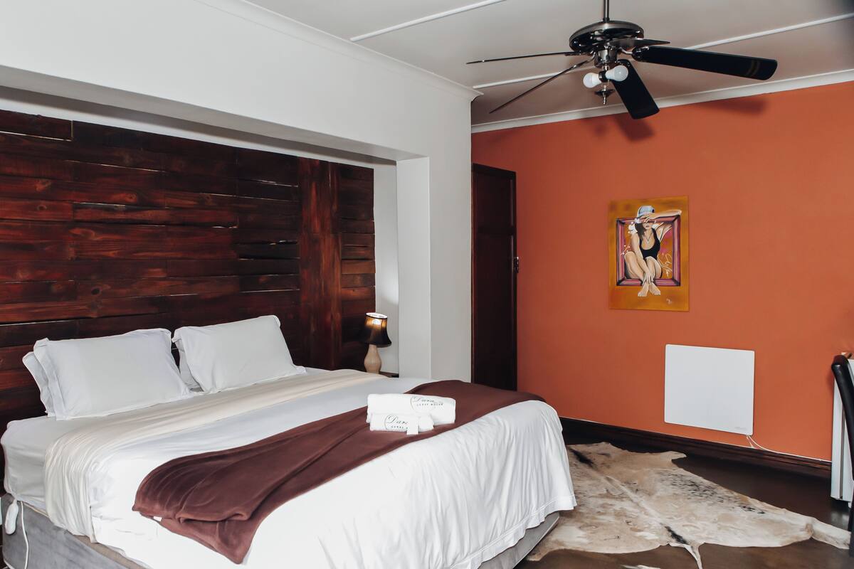 Un dormitorio con cabecera de madera, cama con ropa blanca, manta marrón, ventilador de techo y un cuadro en la pared.