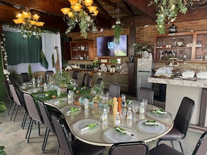 A dining area with a round table set for six, surrounded by chairs, with a brick wall and wooden ceiling in the background.
