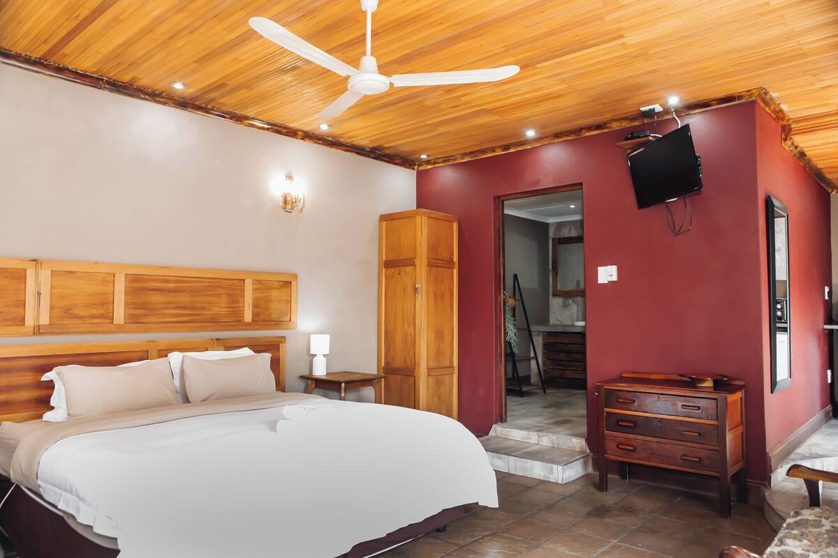 Un dormitorio con cama, cabecera de madera, ventilador de techo, cómoda de madera y televisor montado en la pared.