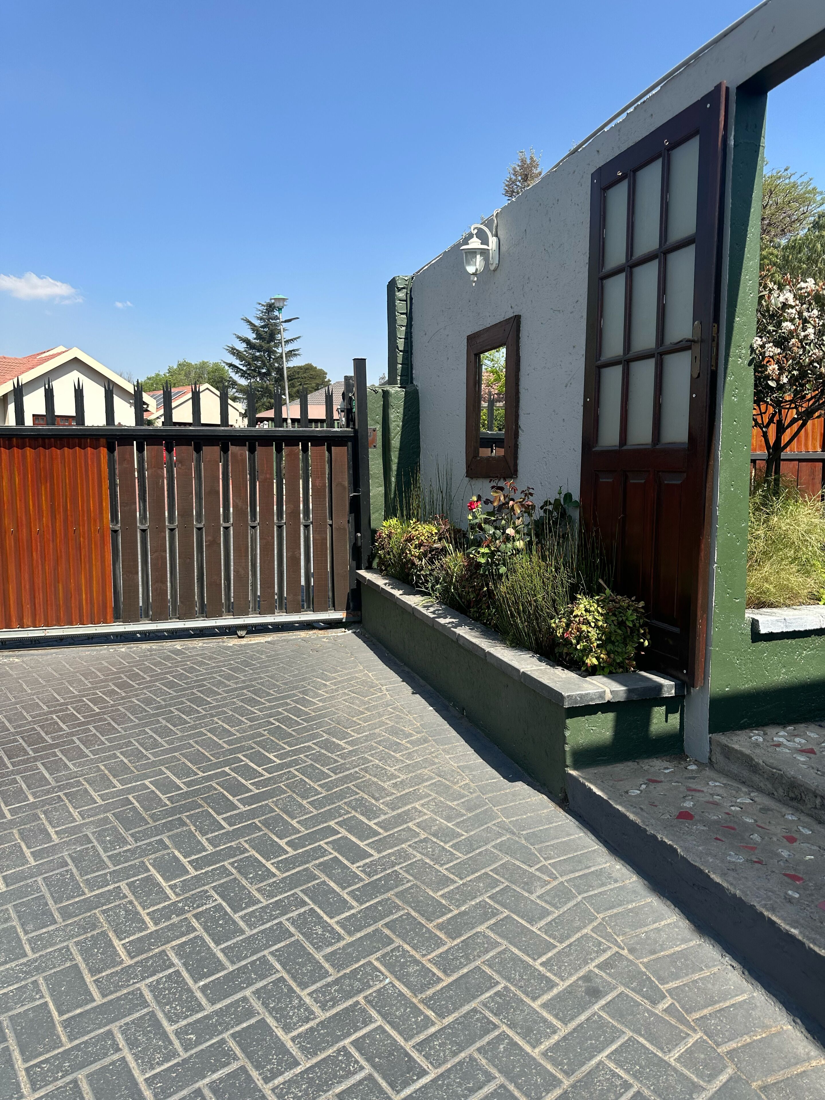 A paved driveway leading to a building with a wooden door and window, surrounded by a fence and greenery.