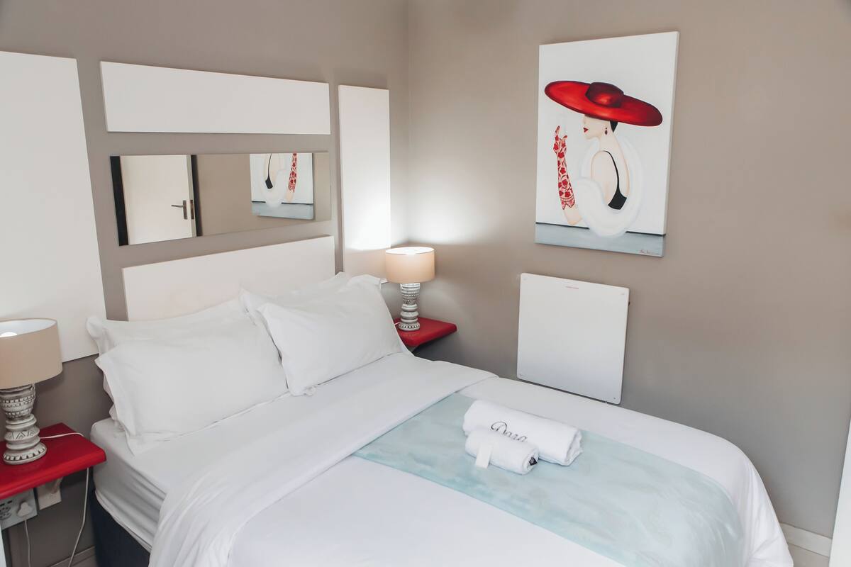 Una cama bien hecha con sábanas blancas, una mesita de noche roja con una lámpara y un espejo montado en la pared.