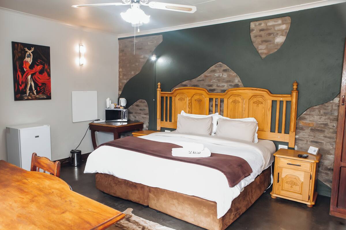 Habitación de hotel ordenada con una cama de madera, un escritorio pequeño, una silla, una mesa de comedor y un cuadro en la pared.