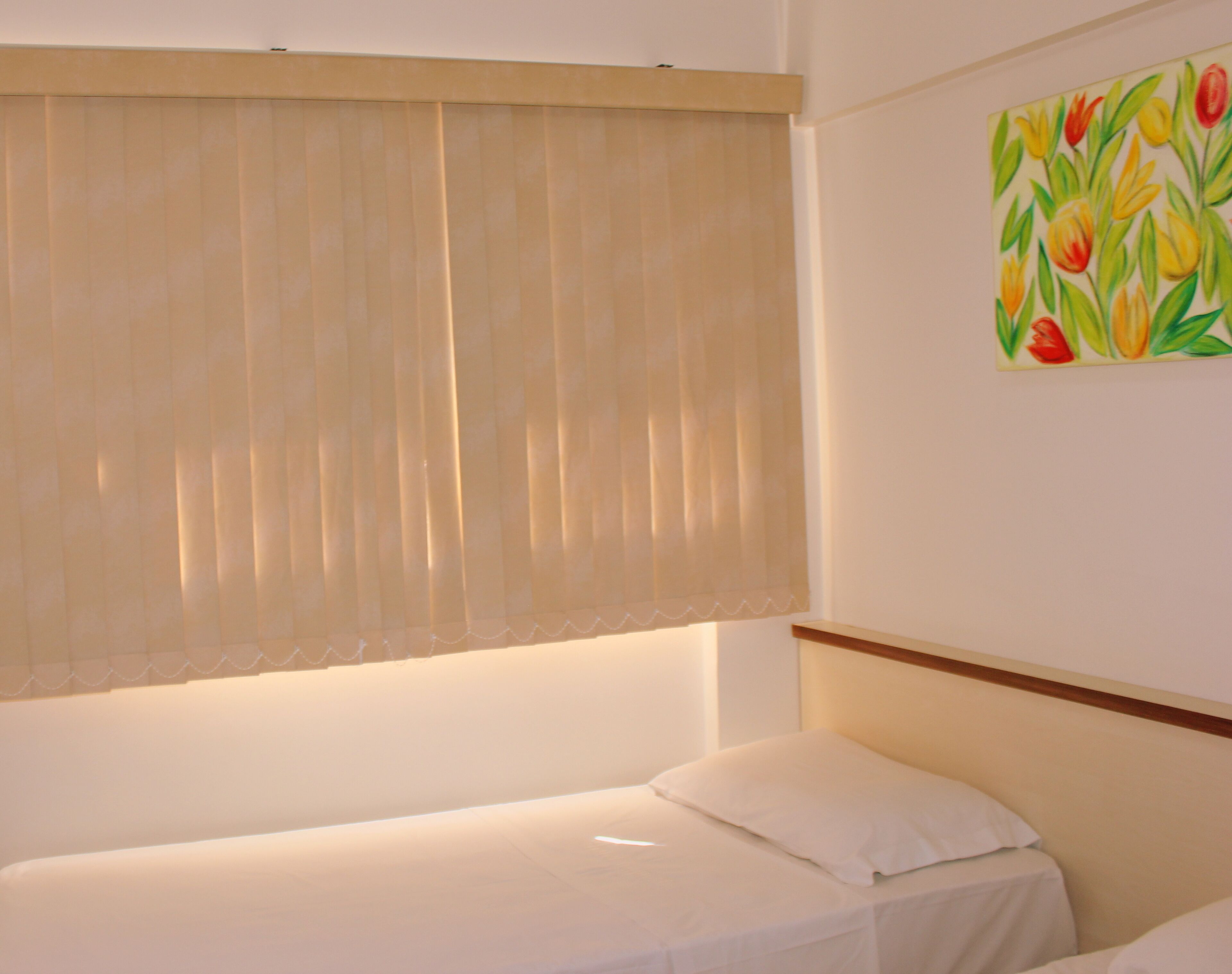 Una cama individual con colchón y almohada blancos, cabecera color beige y ventana con persianas color beige.