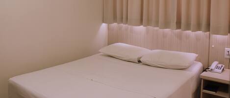 Habitación de hotel con cama, mesita de noche con teléfono y ventana con cortinas.