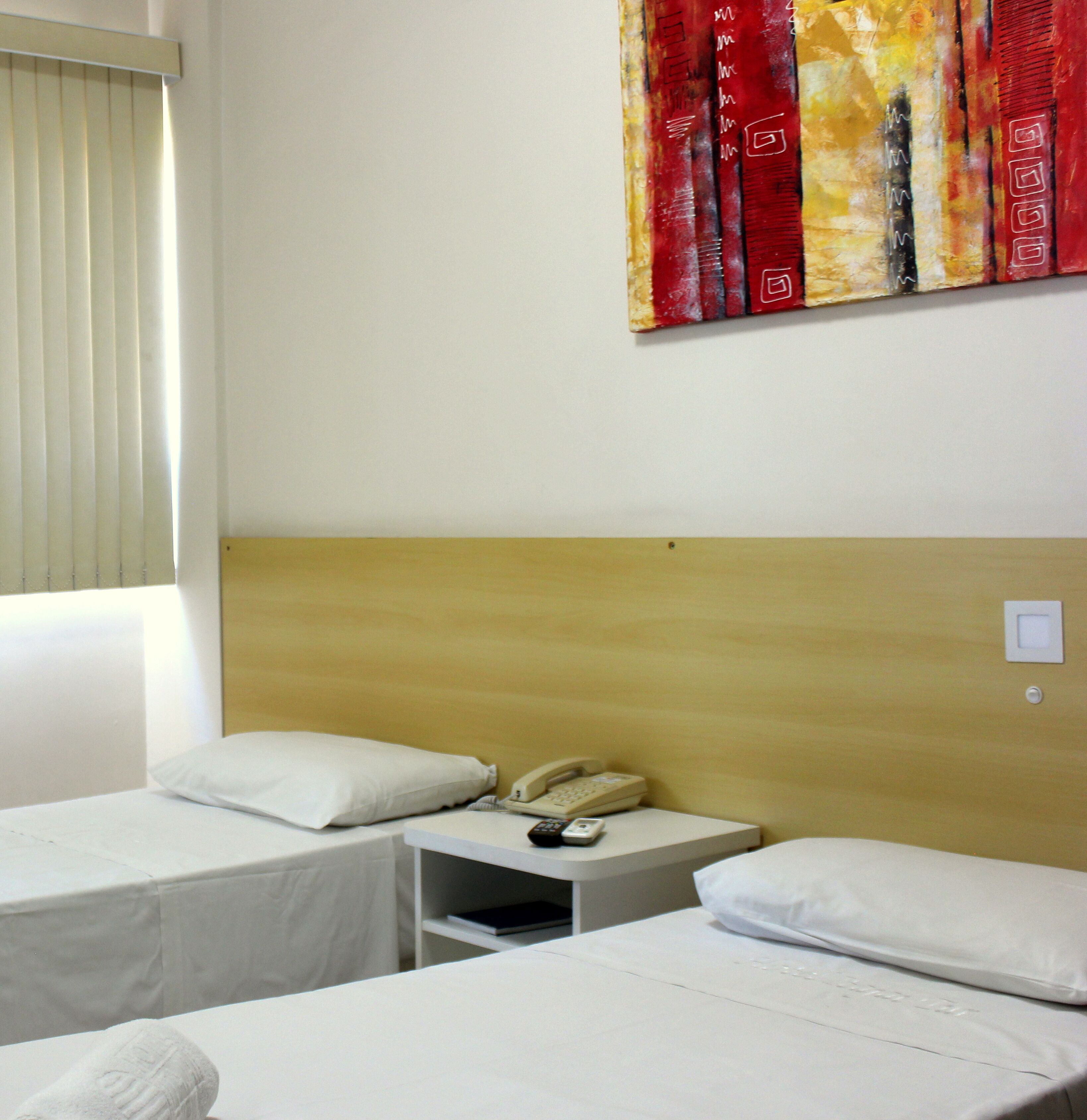 A hotel room with two single beds, a wooden headboard, a bedside table with a telephone, and a window with blinds.
