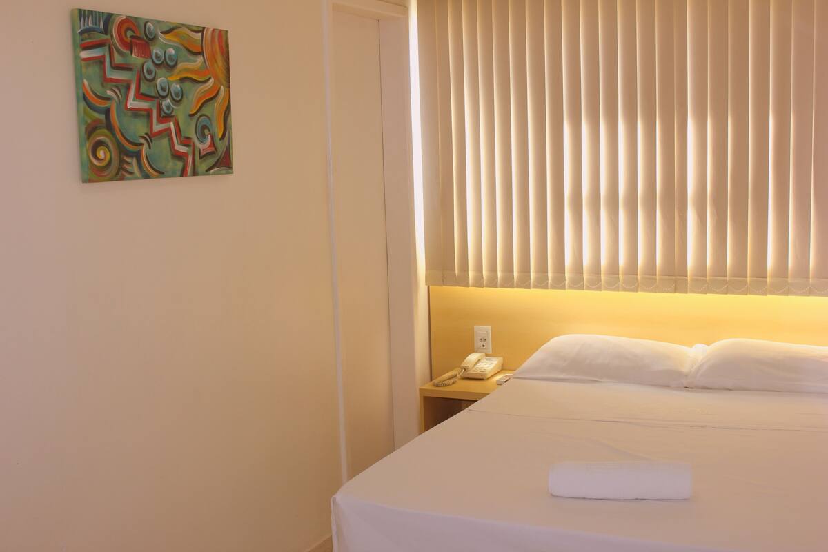 A hotel room with a bed, a bedside table with a phone, and a window with blinds.