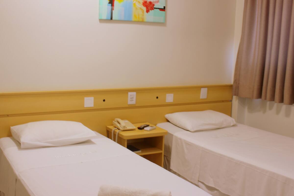 A hotel room with two single beds, a bedside table with a telephone and a book, and a painting on the wall.